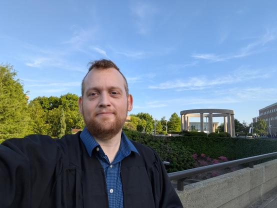 Selfie in graduate regalia in front of a university scene, with trees, buildings, and a colonnade
