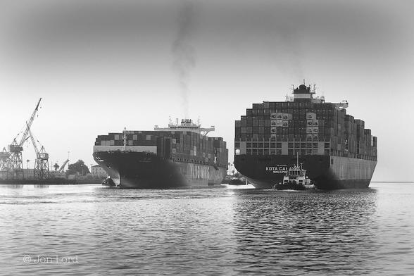 This Is A Black And White Seascape Photo In Landscape Format. The Lower Twenty-Five Per Cent Of The Frame Is Filled With A Body Of Still Dark Water. In The Far Left Are Three Old Cranes On The Waterfront With Their Booms Raised Skywards. To The Left Of Centre Is An Oblique View Of A Large Container Ship Underway And Heading Towards The Camera. The Ship, Like Most Merchant Ships, Has A Dark Hull And White Superstructure. Above Deck Are Multiple Stacked Shipping Containers, Mostly Of A Dark Tone But Some White. A Stream Of Black Smoke Rises Vertically In The Still Morning Air From The Funnel At The Stern. Two Tugs Are Just Visible, One By The Ships Starboard Bow And One At The Stern. The Ships Company Name Is In Large Bold White Letters On The Beam 'YANG MING'. To The Right Of Centre Is A Stern View Of Another Large Container Ship, Roughly The Same Size As The First, Again Under Way But In An Opposite Direction And About Twenty Or Thirty Metres To One Side Of The First. This Second Ship Has Its Decks Stacked With Shipping Containers, A Dark Hull, White Superstructure, A Little Less Dark Smoke From The Funnel And A Single Large Tug In View At The Stern. The Ships Name 'KOTA CAHAYA' Just Visible As Is The Port Of Registry - 'Singapore'. Above This Maritime Scene Is A Clear And Cloudless Sky. 
The Location Is The Los Angeles Main Channel. The First Ship Inbound To The Container Terminal, The Second Outbound To The Pacific Ocean (2018).
