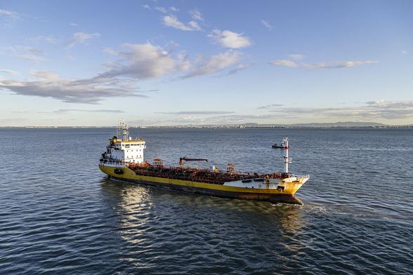 We're looking across calm sea towards low land in the distance with some low hills behind and signs of buildings along the far shore. The main focus is on a ship just ahead of us, pointing to our right and angled slightly in towards us as if in the process of passing us or about to be passed by us. The vessel is a fairly small oil tanker with a long, low deck topped with pipes for the filling and emptying of liquids. The ship is yellow but in need of freshening up as there are heavy signs of rust particularly at the sharp, raised, angled prow. At the aft there is a high bridge structure in white which is gleaming as it's reflecting the low sun of the early evening. The bridge reflects attractively in the dark, rippled water. The sky is mostly clear blue with a few patches of light cloud and the colours emphasise the approach of the end of the day.