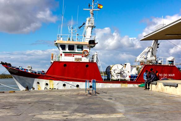 A large ship at the quay in the harbour. The lower part of the photo is a quay. The iron ship lies horizontally on it, a bit higher up. The point at the front is on the far left and the aft deck is on the right of the photo. The back is not quite there, but is behind a building (the fish auction). The cabin is white, but there are two large iron bulkheads on the boat that are painted bright red. One at the cabin and one at the back. The boat is white, with a red border along it. Above it is a beautiful blue sky with white clouds. On the quay on the left, four men are sitting together on a concrete edge of the building. The quay is otherwise empty. A sunny, colourful but also peaceful image.