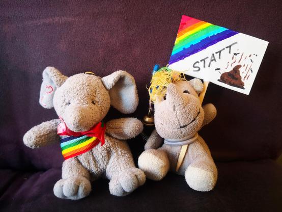 Die beiden kleinen Plüschies Elefanty Elfi, das ihr Regenbogenband schick als Schärpe trägt, und Einhorn Carola mit einem Demoschild vor der Demokratie-Demo am 20.1.2024, im Hintergrund der Stoff (Sitzfläche und Rückenlehne) des dunkelvioletten Sofas.