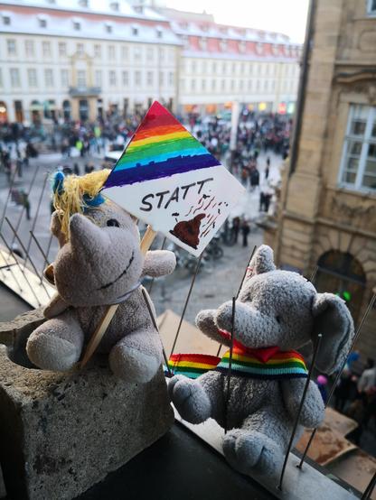 Die beiden kleinen Plüschies Elefanty Elfi, das ihr Regenbogenband schick als Schärpe trägt, und Einhorn Carola mit einem Demoschild nach der Demokratie-Demo am 20.1.2024 auf der Wohnzimmer-Fensterbank, im Hintergrund die sich langsam auflösende Demo auf dem Maxplatz.