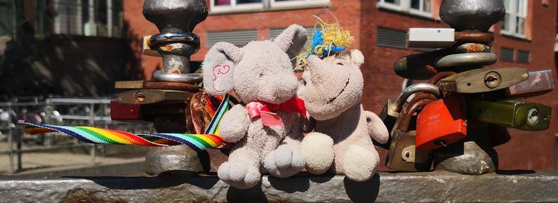 Elfi und Carola aneinandergeschmiegt auf dem Geländer einer Brücke, Elfis Regenbogenband flattert im Wind.