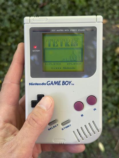 A person holds a classic Nintendo Game Boy displaying the title screen for “Tetris,” which reads: “TETRIS™,” with options “1 PLAYER” and “2 PLAYER” and the text “©1989 Nintendo” below.