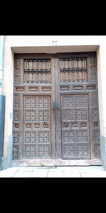 An old and weathered wooden set if doors with natal studs with a set of metal grill panels at the top and the number 77