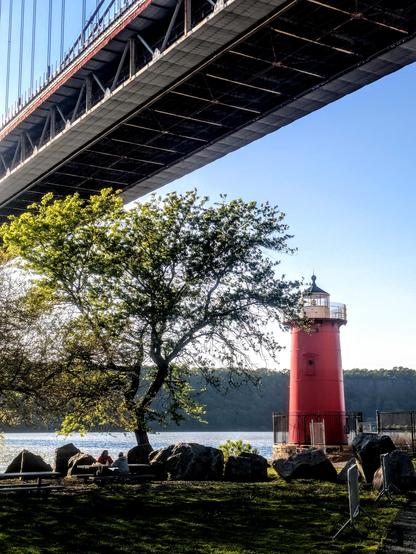 Golden hour. A small bright red lighthouse on the bank of a river. Near it is a tree, under which two young men are sitting at a picnic table. Partially seen above is a very large suspension bridge.