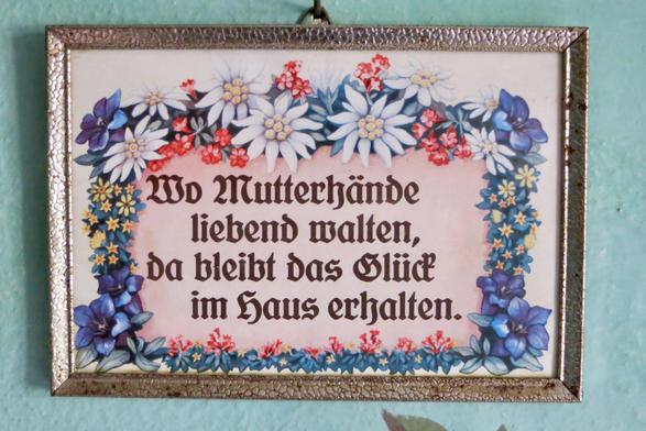 Sinnspruch in Frakturschrift und von einer Blumenborte umrandet: Wo Mutterhände liebend walten, da bleibt das Glück im Haus erhalten,