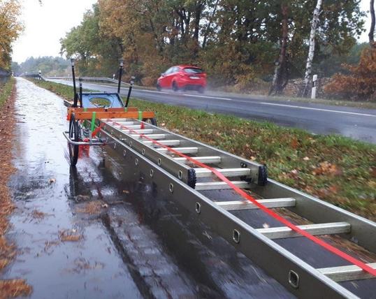 Ein Fahrradanhänger steht an einer regennassen Straße. Darauf befestigt ist eine lange, ausgezogene Leiter.