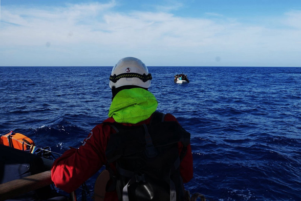 Die NADIR nähert sich einem überfüllten treibenden Schlauchboot. Von hinten ist ein*e Seenotretter*in mit Schutzhelm zu sehen, die hinüber blickt.