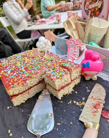 Kuchen auf Schieferplatte, im Hintergrund ein Tablett mit Holzbesteck, einer Candy "Maschine" und Pappbechern, sowie Kinder an Tischen im Hof mit pinken Trinkbechern und Tellern. Der Kuchen ist in der Mitte mit Himbeermarmelade gefüllt, obendrauf ist eine pinke Himbeermarmeladenzuckergussglasur und bunte Zuckerperlen, -streusel und -sterne. Der Kuchen ist zur Hälfte gegessen, einige Stücke sind schon geschnitten. Eine My Melodie-Figur, ein großes Messer und ein Tortenheber liegen daneben.