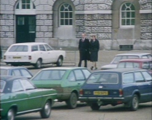 A street scene from the show. Two characters are seen from a distance walking on a sidewalk as if shot from an upstairs window across the street, while traffic is shown in the foreground, in this case a group of then-nondescript but now-rare cars from the 1970s.