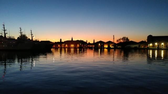 Über eine lange Wasserfläche hinweg sind in mittlerer Ferne Gebäude zu sehen, hinter denen gerade die Sonne untergegangen ist und sie mit einer Farbaura umgibt. Die flachen Gebäudefronten wiederum sind in Teilen von wie Kerzen wirkenden Leuchten erhellt. Tiefblaue und Orangetöne sind bestimmend im Bild. Am linken Bildrand befinden sich einige größere Schiffe, fast nur als Umriss zu sehen, deren Anlagenmasten in den Himmel ragen.