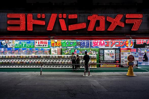 Silhouettes of a couple with their child standing in front of a long row of capsule toys vending machines in Shinjuku in Tokyo at night. The vending machines are standing in front of an illuminated department store topped with a giant black sign with the store names in big red neon letters.