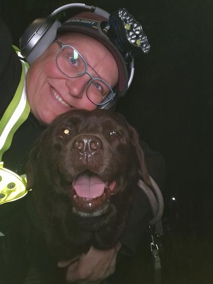 Night time selfie of a person and a chocolate brown Labrador with greying snout. The person is wearing a black jacket, yellow safety harness, red cap, silver headphones and blue-rimmed glasses. On their cap rests a headstrap torch in off position. Their free hand is around the dog's shoulders. Both are facing the camera. The dog's mouth is slightly open, showing off some mlem. The person's head is resting on the dog's head. They're smiling, grateful for their time spent together.