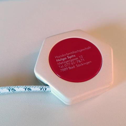 A small measuring tape in a white hexagonal plastic case with a red circle in which the address of a specialised needlework shop is printed.