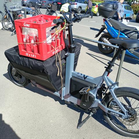 Ein hellviolettes Lastenrad auf dessen vorderen abgedeckten Transportkorb eine leere Kiste Cola verzurrt ist.