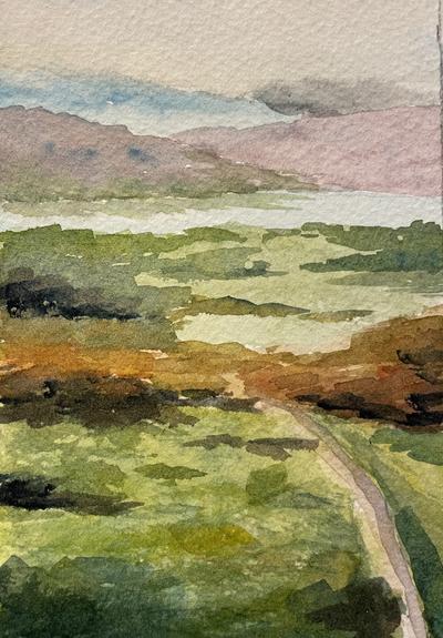 A winding path cuts through a patchwork of textured fields and brush, leading toward distant hills that rise gently beyond a quiet body of water.