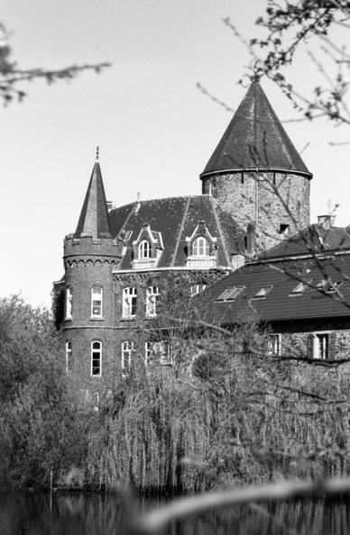 Black and white photograph of a picturesque, historic castle with conical turrets and steeply pitched roofs, partially obscured by leafless tree branches. Part of the building is reflected in a calm body of water in the foreground