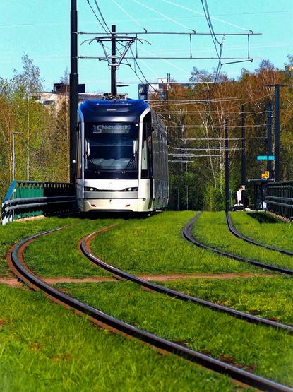A modern tram approaches on a curved track surrounded by green grass. The tram is labeled "15 Kageludden (M)" and features overhead electrical lines. In the background, there are trees and a faint urban landscape.