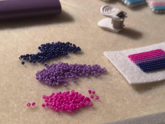 A bead mat with three piles of beads in the bisexual pride colors and a piece of pellon with a beaded bisexual pride flag. The corner of a trans pride flag pin can be seen in the corner. You can also see a purple lighter and a little white bead stopper clip.