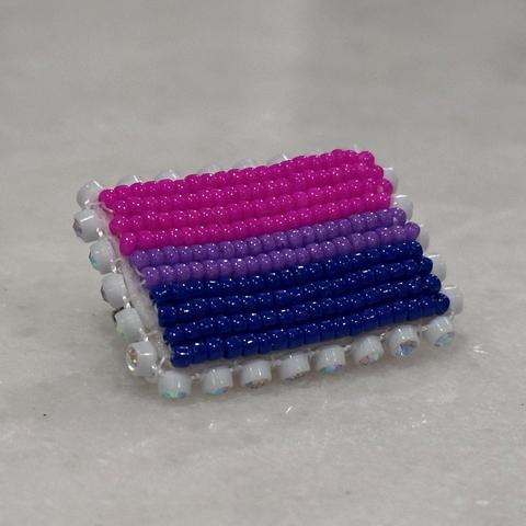 A beaded bisexual pride flag pin edged with rhinestone banding on a white marble background