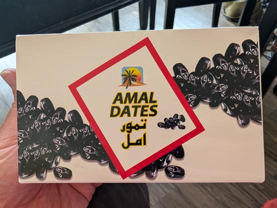 A box of Amal Dates. The box has pictures of smallish black dates and text in Arabic.