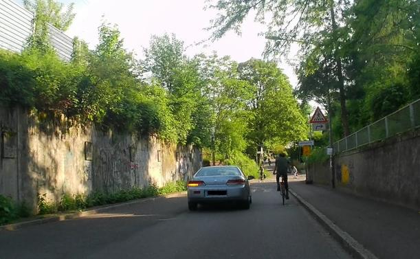 Sicht auf alten Sportwagen und jugendlichen Fahrradfahrer von hinten. Die hochbetagte Fahrerin überholt mit Unterschreiten des Mindestabstand um ca. 80 cm.