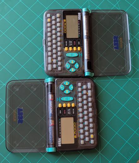 Both of the Sega pdas side by side.

The front of the PDA. The screen is on and displaying icons like a phone book, world clock calculator,  IR communicator etc. 

The shell is a translucent gray. With various buttons in Gray, yellow and turquoise.