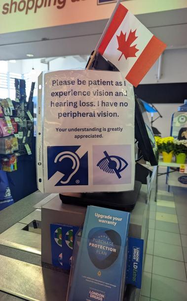 A customer service point-of-sale area shows a printed sign in a plastic sleeve with the text "Please be patient as I experience vision and hearing loss. I have no peripheral vision. Your understanding is greatly appreciated." Below the text are graphic symbols representing hearing impairment and vision impairment. A small Canadian flag is stuck into the top of the POS terminal. To its right is a screen and a display of yellow-flowered plants. Next to the sign is a display of a blue “PURCHASE PROTECTION PLAN” warranty card and a blue London Drugs "LD Extras" rewards program card.