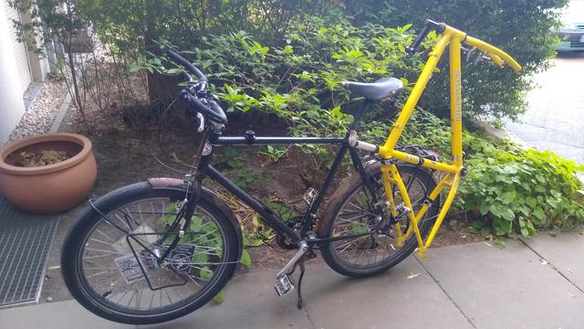 Ein schwarzes Fahrrad. An den Gepäckträger ist seitlich ein gelber Fahrradrahmen mit dem hinteren Rahmendreieck so  festgezurrt, dass die Sattelstütze (ohne Sattel) nach vorne zeigt und die Gabel nach hinten