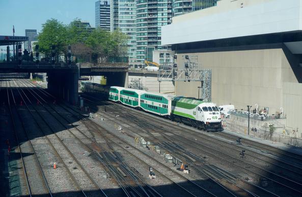 Photo of a rail corridor in a built-up urban area, looking along and a little across the corridor. A train is in the centre of the photo. Tall buildings are seen immediately next to the tracks. In middle distance, a few trees are seen growing in a little parkette built on a bridge span above the tracks.

The corridor has eight tracks visible in the photo, with some crossover tracks between them. It is divided into two sets of four tracks by a long pillar holding up the bridge. There is no electrification. One short signal gantry is seen over the two farthest tracks, and ground-level signal heads are between other tracks.

The train is on the track farthest from the camera, and is heading towards to the right side of the photo. It is led by a locomotive and has six passenger cars. The cars are bilevel, as seen by two rows of windows. The paint scheme is green and white, two different shades of green are used on the locomotive, and a different shade is on the passenger cars.

On the right side of the photo is a blank concrete wall of a building in foreground, and beyond that greenish grass of residential towers. At the left edge of the photo, an awning above the bridge has the Marriott hotel logo. The bridge itself is significantly wider than needed for just a street, and creates a little parkette above the tracks. The trees are maybe about 6 metres tall and in spring leaf. A bit of the blue sky is visible in top left, it is sunny and the shadows in the photo are deep.