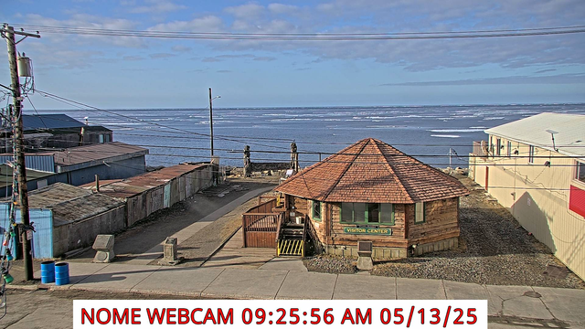 Webcam image from 925am Tuesday, May 2023 looking south across Front Street in Nome. A few buildings are visible and the Bering Sea beyond with scattered decaying sea ice. Some mid-level clouds are in the sky above the ocean.