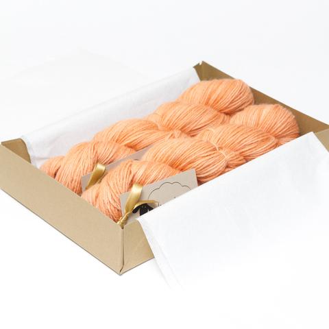 Picture shows an open A4 box inlaid with white tissue paper containing two skeins of yarn in shades of orange.