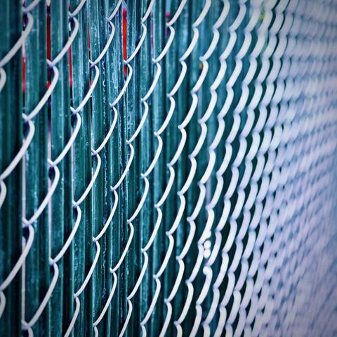 A close-up perspective view of a silver chain link fence reveals vertical green slats down the length of it into the blurry distance.