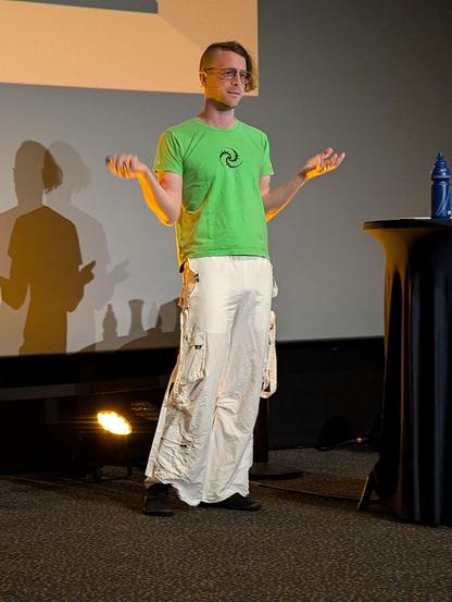 Oli-Obk presenting, wearing white cargo pants and a green t shirt, arms with palms facing up, as if shrugging