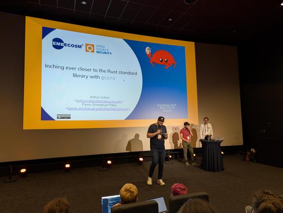 A big screen with a slide with the title "Inching closer to the Rust standard library in gccrs". On the bottom right, the presenters and MC.
