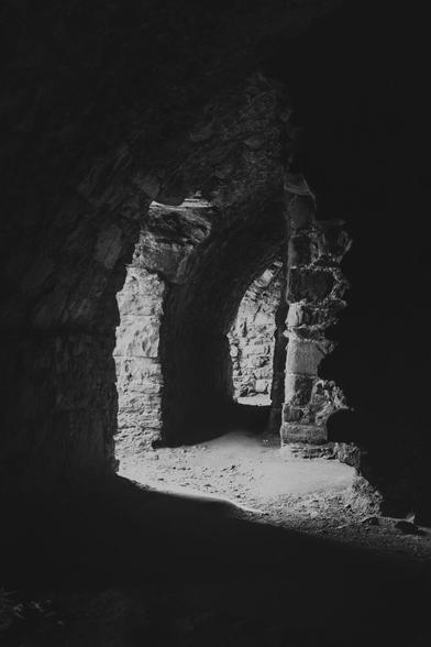 Schwarzweißfoto mit Kellergewölbe im Halbdunkeln. Etwas Licht scheint an einer Stelle links im Bild von draußen hinein.