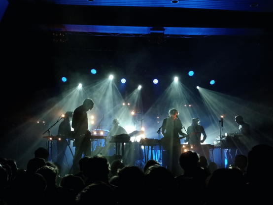 A concert hall shot from the audience with many musicians on the stage, in contrast-rich rather monochrome blue-green lighting against a black (unlit) backdrop. Each of the six musicians has their own white-ish light cone from above, most with their table based electronic instruments, one with an electric guitar.