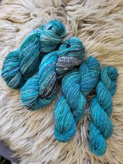 Four hanks of hand-spun yarn lie on a sheepskin. The two strands on the left are predominantly bluish-green with black and white (salt&vinegar) and purple sections. The two strands on the right are a more uniform bluish-green colour.