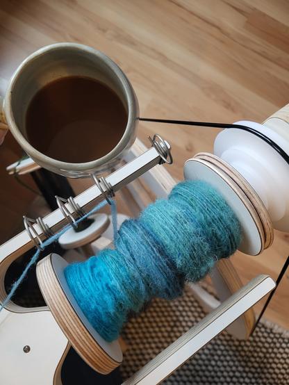 Close up of the bobbin on my spinning wheel. There's a single thread in blue/green/teal tones on it. Next to it, there's a cup of coffee.
