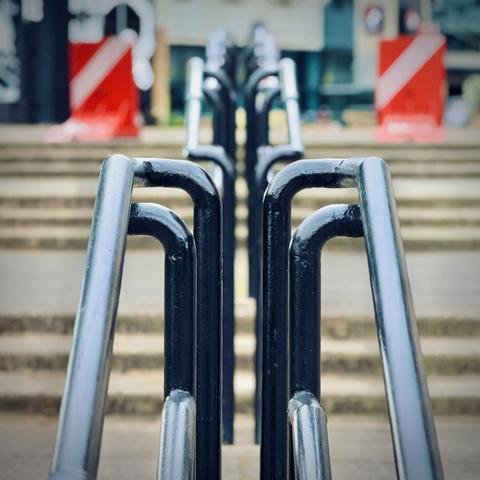 Four navy-blue handrails up four separate three-sets of outdoor concrete stairs are viewed straight up the line, with an orange barricade on each of the two sides at the top of the sets in the blurry distance under an overcast sky.