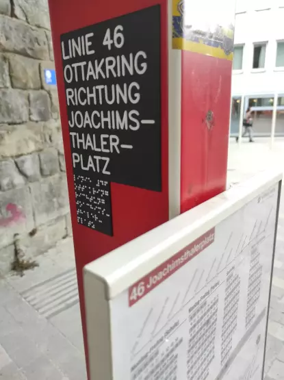 Auf einem roten, rechteckigen Masten mit den Fahrtzeiten der Linie 46 befindet sich ein schwarzes Schild auf dem in weißer Profilschrift und Braille „Linie 46, Ottakring, Richtung Joachimsthalerplatz“ steht. Dahinter befindet sich ein Blindenleitsystem, eine Mauer und ein weißes Gebäude mit großen Schaufenstern.