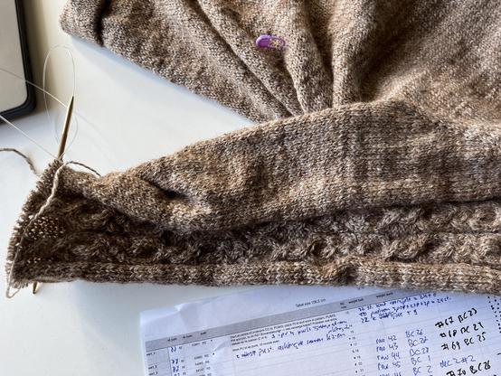 Sweater sleeve with a cable panel on bamboo needles lying on a white table with some notes to keep track of the knitting.