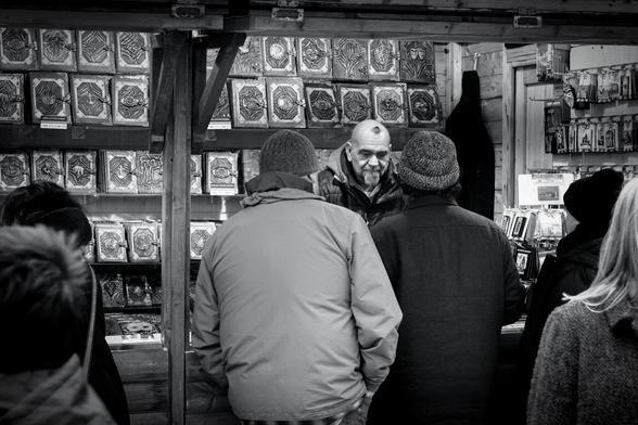 A man is working behind the counter of a craft stall at the Christmas Market at London's Trafalgar Square. He has a grey goatee beard and a small wisp of dark hair looking somewhat like a spider, or perhaps a small hairy Vesuvius on his shaved head.  He looks sad as he serves a man and a woman while others look at the goods on his stall. As well as fridge magnets and souvenirs, the main thing he's offering are leather-bound journals in three sizes, with some kind of decorative metalwork on the covers, some of which contains masks, faces, skeletal hands, anchors, ampersands, and elephants, along with many more abstract designs.