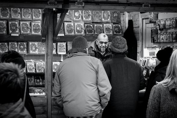 A man is working behind the counter of a craft stall at the Christmas Market at London's Trafalgar Square. He has a grey goatee beard and a small wisp of dark hair looking somewhat like a spider, or perhaps a small hairy Vesuvius on his shaved head.  He looks sad as he serves a man and a woman while others look at the goods on his stall. As well as fridge magnets and souvenirs, the main thing he's offering are leather-bound journals in three sizes, with some kind of decorative metalwork on the covers, some of which contains masks, faces, skeletal hands, anchors, ampersands, and elephants, along with many more abstract designs.