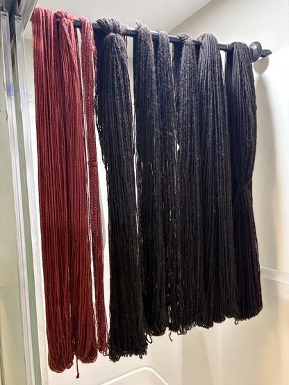 Skeins of yarn, hanging loose from a walking stick over the top of a bathtub shower. The three closest the viewers are red or red-pink, followed by six black (very dark brown) ones. Some of the black ones are quite large, looking all squishy. Don’t squish them, though, unless you want to get wet.