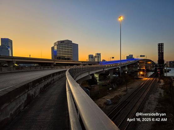 Early morning view along the pedestrian crossing of a large highway bridge, next to an old railroad train trestle and drawbridge, with the downtown skyline and Saint Johns River in the background as warm, glowing colors rise from the horizon.