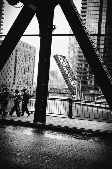 A black-and-white photograph depicting a bridge with a raised section, surrounded by modern buildings. People walk along a wet street in the foreground, with reflections on the pavement. The scene conveys a urban atmosphere.