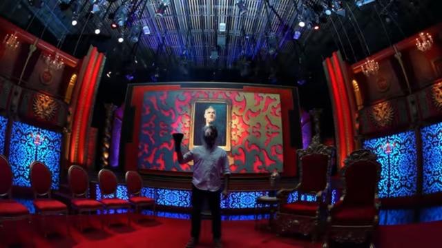 A brightly lit stage set features a man holding a microphone in front of an ornate backdrop displaying a large portrait. The scene includes elegant red and blue decorations, with empty red chairs arranged in front.