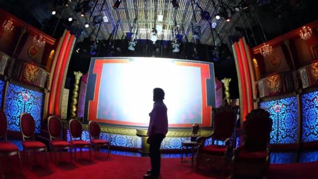 A stage setup with vibrant red and blue decor, featuring ornate chairs and a large illuminated screen. A person stands silhouetted in the foreground, facing the screen.